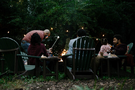 Black friends gathering around fire-pit roasting marshmallows and eating smores having a good time