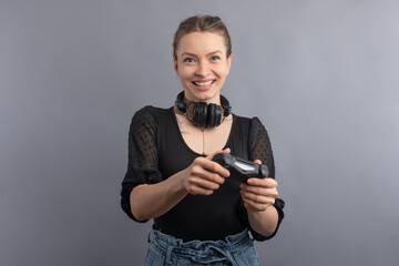 Beautiful smiling woman with pigtails and emotional face using joystick and headphone on gray background. Concept of playing video game. Active entertainment. Modern technology.
