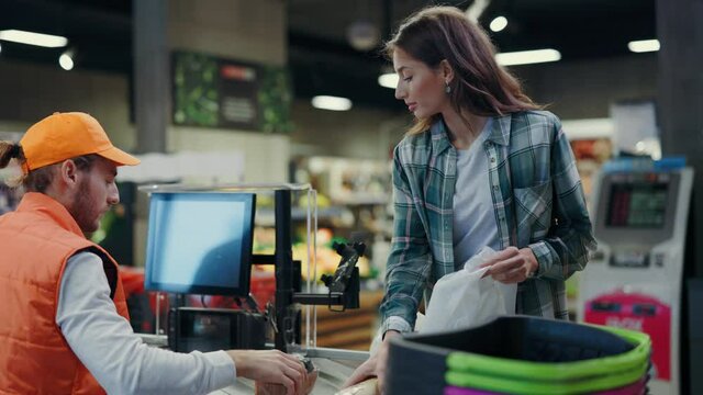 Caucasian woman talkking with cashier bearded man scannin food items and telling price. Customers. Consumerism. Supermarket services. Communication.