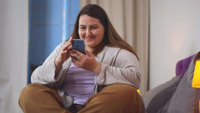 Portrait of obese smiling woman sitting cross-legged on bed and chatting on smartphone