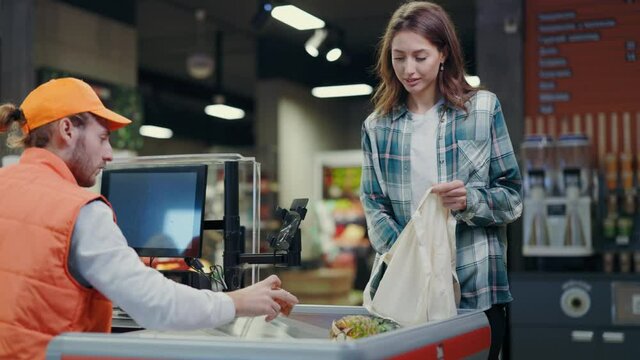 Young beautiful woman standing at cash register communicating with cashier packing her products in plastic bags. Supermarket. Grocery store shopping.