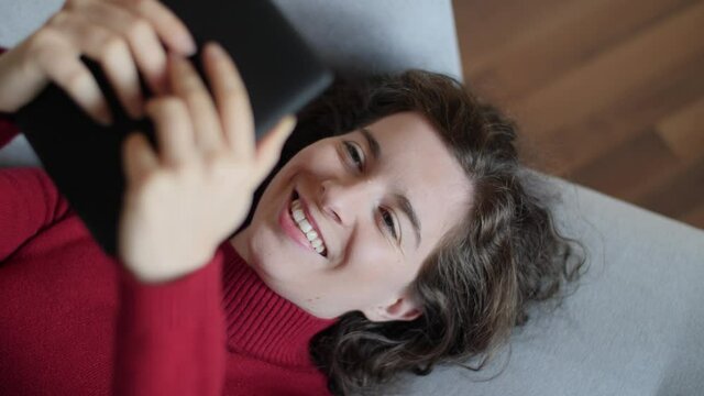 Young woman with red pullover reading e-book on grey couch at home. Beautiful caucasian girl lying on cozy sofa looking at modern e-book reader. Slow-motion, close-up, natural daylight.