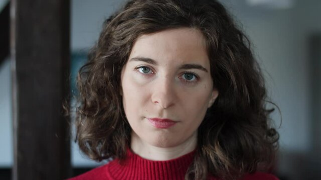 Portrait of beautiful confident young woman opening eyes and looking at camera. Attractive millennial girl with curly hair, red pullover and natural make-up at home indoors. Slow-motion.