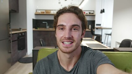 Smiling young man making selfie or video call while sitting at home on sofa. Portrait of male blogger doing a video chat from his apartment. - Powered by Adobe