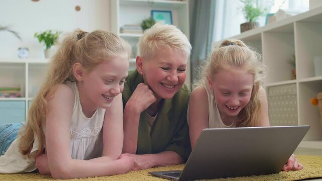 Medium close-up of white-haired Caucasian woman laying down on floor with two blue-eyed little granddaughters. Grandma watching video on laptop, talking, laughing with kids