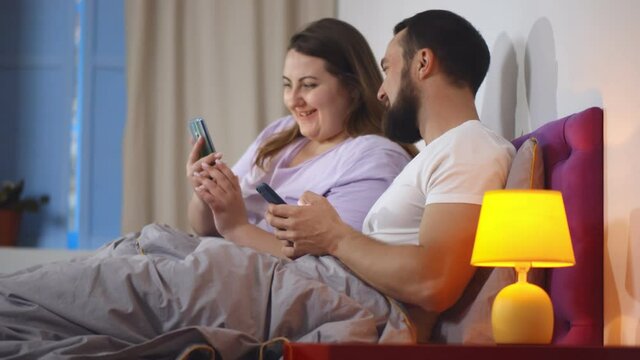 Loving young couple holding mobile phone watching photos or videos together in bed