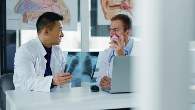 Caucasian and chinese young adult doctors discussing lung disease with x-ray image negotiating collaborating inside private office medical healthcare hospital.