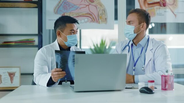 Multi-race young doctor couple negotiating in medical office. Chief asian therapist talking to assistant discussing lung disease xray image coronavirus infection.