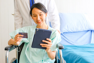 Happy Female Asian patient hold a credit card and Tablet while sitting in wheelchair in hospital...