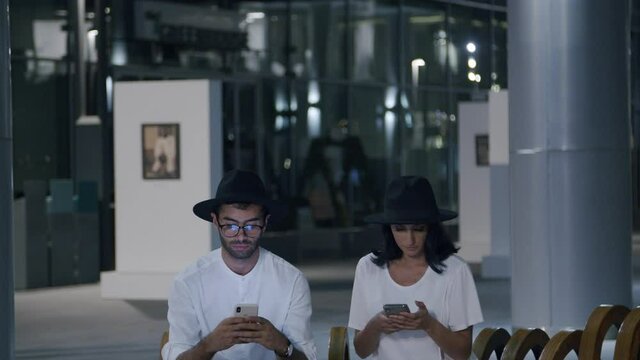 Man And Woman Wearing Black Hats Texting On Their Mobile Phones While Sitting Inside The Building Hallway - Medium Shot