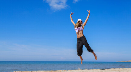Obraz premium New year's eve on the beach by the sea. happy girl in a Santa hat in a jump. Journey to Christmas. Holidays in hot countries. hair like a beard