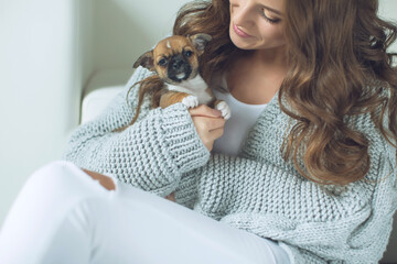 The owner of the dog and her pet. Happy ethnic girl holding a cute puppy near her face, expresses love and care for a pet, woman in a sweater. High quality photo.