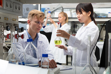 Fototapeta premium Positive Chinese female working with reagent in test tube during chemical experiment with male fellow student in university lab. Focus on male