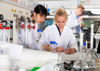 Two female students of Faculty of Chemistry performing experiments in university laboratory, recording results in workbook