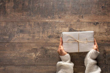 Top view on young woman's hands holding Christmas, birthday or any other celebration gift wrapped in white paper on rustic wooden background. Preparing for holidays and celebration. - Powered by Adobe