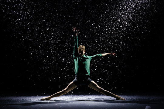 Silhouette of a man performing modern choreography or contemporary ballet against the background of falling snow in the rays of studio light.