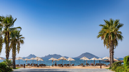 Umbrellas and sunbeds on beautiful beach with palm trees and islands on horizon in Mediterranean sea. 