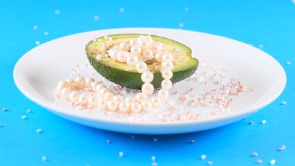 half a fresh avocado with pearls on a white plate covered with rhinestones on a light blue background.