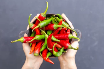 Gardinen Chilischoten Top view of female hands holding green and red hot chili peppers in a rusty bowl on a dark tabletop  © Neven Krcmarek/Wirestock