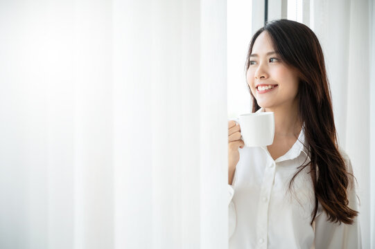 Young asian beautiful woman clothing white pyjamas and drink coffee beside window at morning in bedroom. She feeling happy fresh and smile relax