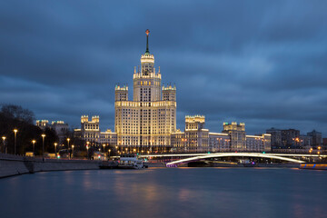 Night cityscape of brightly lighted empire style skyscraper on a riverbank. Bright bridge in front...