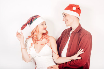 Obraz premium Couple on a white background in santa hats. The girl and the guy are smiling, new year, christmas. Woman in a beautiful white dress and pink hair, in accessories