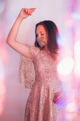 Naklejka premium Christmas festive look. Pandemic fashion. Ethnic style. Beautiful woman in east glossy dress designer golden chain crystal beads face mask posing in pastel neon light blur lens flare.