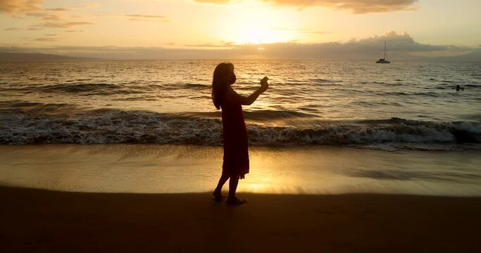 Silhouette of a long-haired woman wearing a dress taking photos with her cell phone at sunset on a tropical beach. Waves are rolling in from the calm ocean, and they are rolling across her feet.