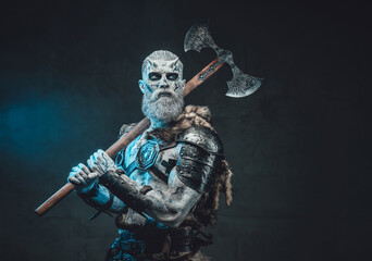 Evil fashion of king of the dead with naked torso and pale skin in dark background with two handed axe on his shoulder.