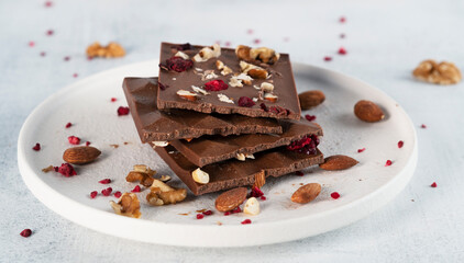 Broken dark healthy chocolate bar with nuts and freeze dried raspberries on white plate. Tasty food. Side view. Horizontal orientation.