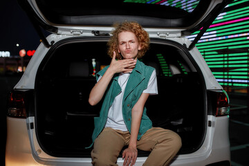 A cool-looking redhead young woman with freckles looking into a camera sitting and posing inside of an opened car trunk outside on a parking site with a led screen behind