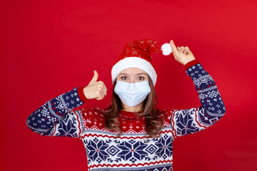 a young woman in a medical mask, Santa Claus hat raises her thumb up, isolated on a red background with space for text.