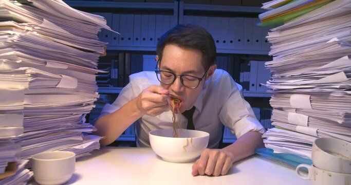 Hungry asian business man eating instant noodles for a late meal while working hard at late night. Exhausted overload alone male take off glasses and lay down on desk covered with stack of paperwork.