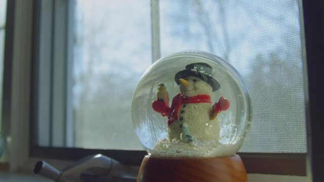 Snowflakes are settling in a snow globe as it sits in a window during the day