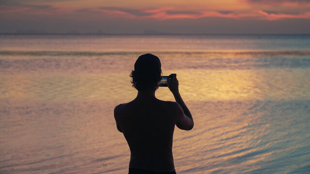 Athletic man standing on the beach at sunset and takes pictures on smartphone