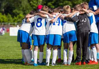 Coach with children soccer team. Kids in white and blue soccer uniforms huddling in a team. Boys motivating in a circle before the tournament game. Football match for youth teams on school pitch