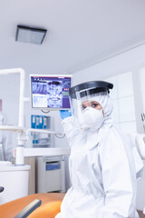 Patient pov in stomatological office listening doctor talking about teeth treatment showing x-ray. Stomatology specialist wearing protective suit against infection with coronavirus pointing at