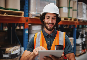 Cheerful young caucasian smiling worker with helmet on head using tablet for work while standing in warehouse against goods