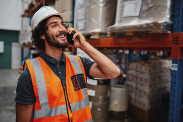 Happy bearded warehouse manager in conversation on mobile phone wearing white helmet and safety...
