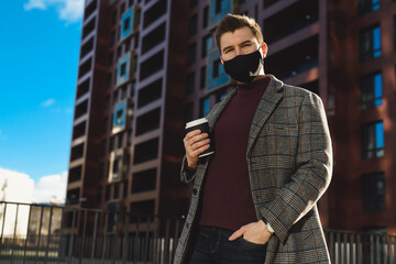 Portrait of a young businessman or manager with a cup of coffee. Social distancing in a big city during coronavirus Covid-19 pandemic. Wearing a mask in a public place.