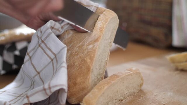 A slice of white bread being cut from a bread loaf with a knife on a wooden cutting board while another slice of bread is laying beside. Slow motion with shallow depth of field.