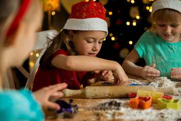 Mother and little kids in red hats cooking gingerbread cookies and playing. Beautiful living room with lights and Christmas tree, table with lantern. Happy family celebrating holiday together.