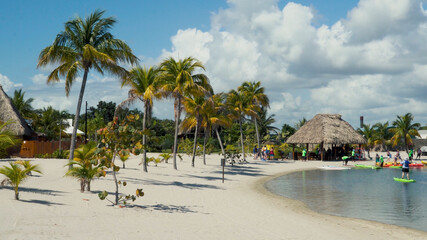 Beautiful beach in Belize. Palm trees, white sand, thatch roof building, tourists relaxing and doing water sports. People in the shot are distant and unrecognizable, beautiful sunny day