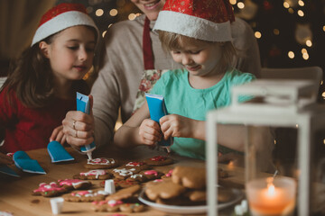 Mother and little kids in red hats cooking gingerbread cookies and decorating with glaze. Beautiful living room with lights and Christmas tree. Happy family celebrating holiday together.