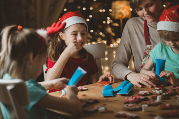 Mother and little kids in red hats cooking gingerbread cookies and decorating with glaze. Beautiful...