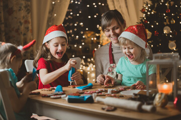 Mother and little kids in red hats cooking gingerbread cookies and decorating with glaze. Beautiful living room with lights and Christmas tree. Happy family celebrating holiday together.