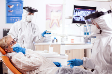 Fototapeta premium Senior patient presenting teeth pain wearing hazmat suit agains coronavirus at dentist. Elderly woman in protective uniform during medical examination in dental clinic.