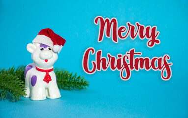 Happy Christmas bull or cow toy in red Santa hat and red bell on the neck with green fir branches on blue background as symbol of 2021 year. Greeting card with Merry Christmas words.