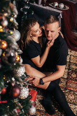Attractive couple at Christmas at home near a Christmas tree hugging each other. New Year's photo of a guy and a girl in a New Year's interior, a cozy decorated house.
