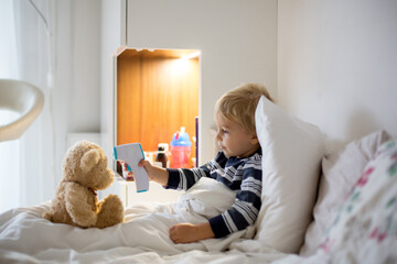 Little toddler blond child, playing with teddy bear in bed, while being sick, checking his temperature